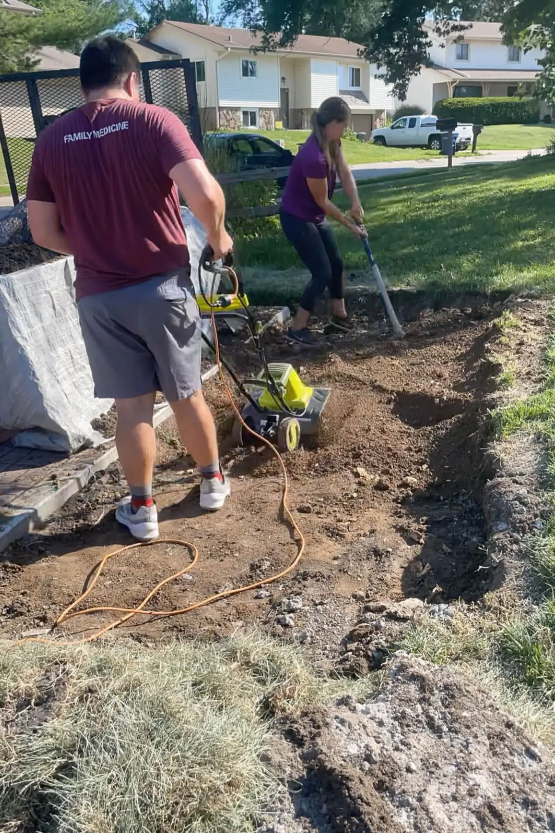 Couple working together to level and compact the dirt base using a pick axe and electric tiller for a DIY paver pad install.